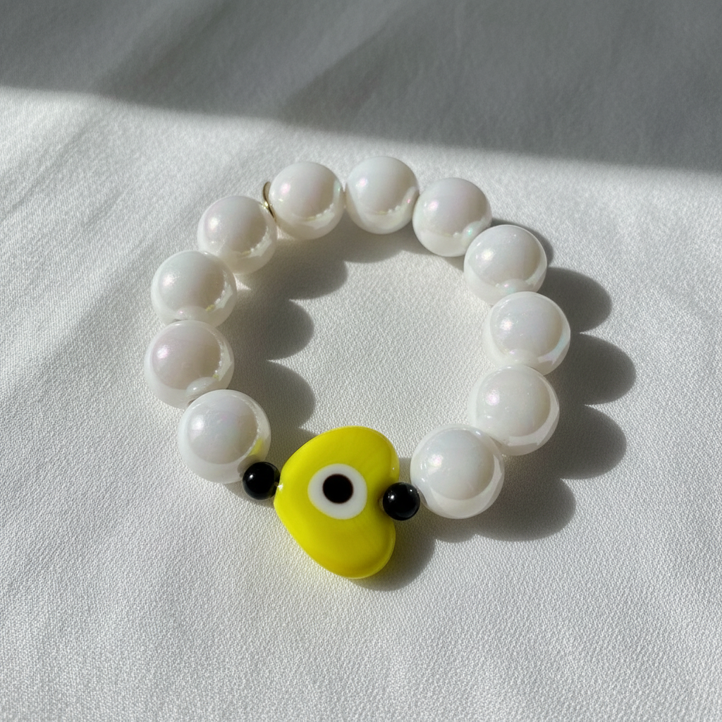 The Aegis Sol Heart Bracelet - White beaded bracelet with a yellow eye guardian heart charm on a white fabric background