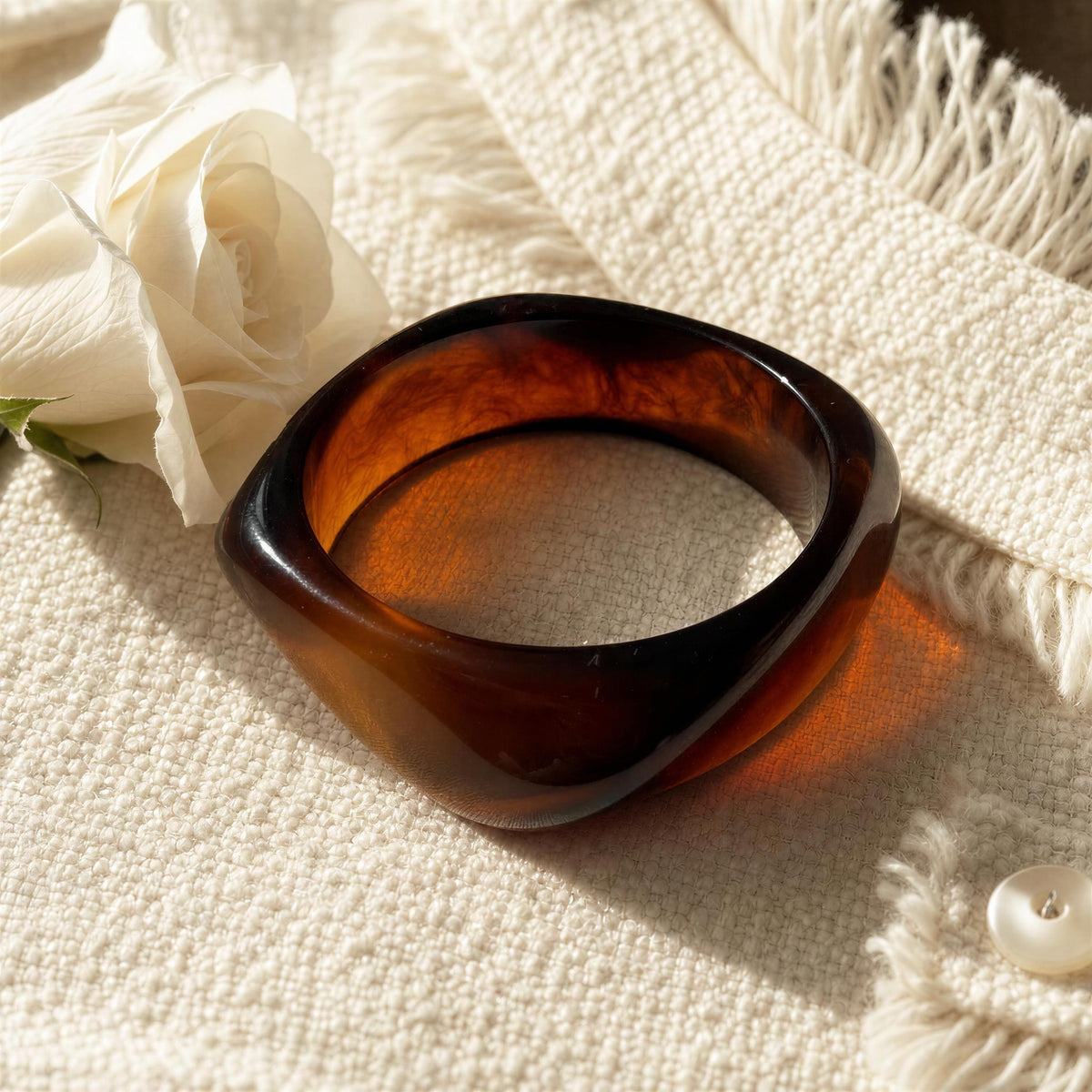 A thick, square-shaped tortoiseshell bangle bracelet with a glossy finish, resting on a textured cream-colored knit blanket next to a white rose.