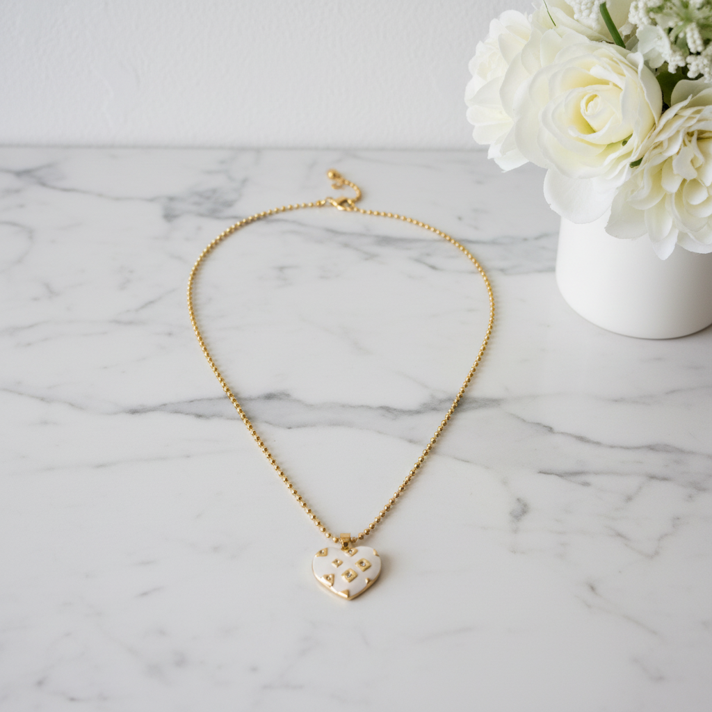 Gold necklace with a heart-shaped pendant on a marble surface with white flowers.