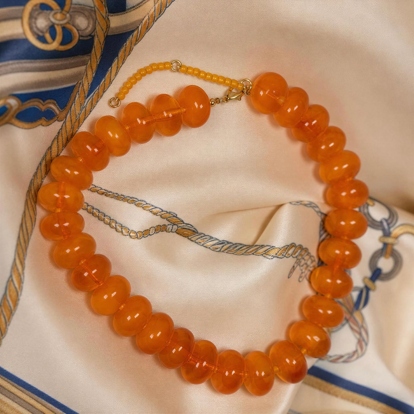An orange beaded necklace with a gold clasp, arranged on a cream-colored silk scarf with blue and gold rope patterns.