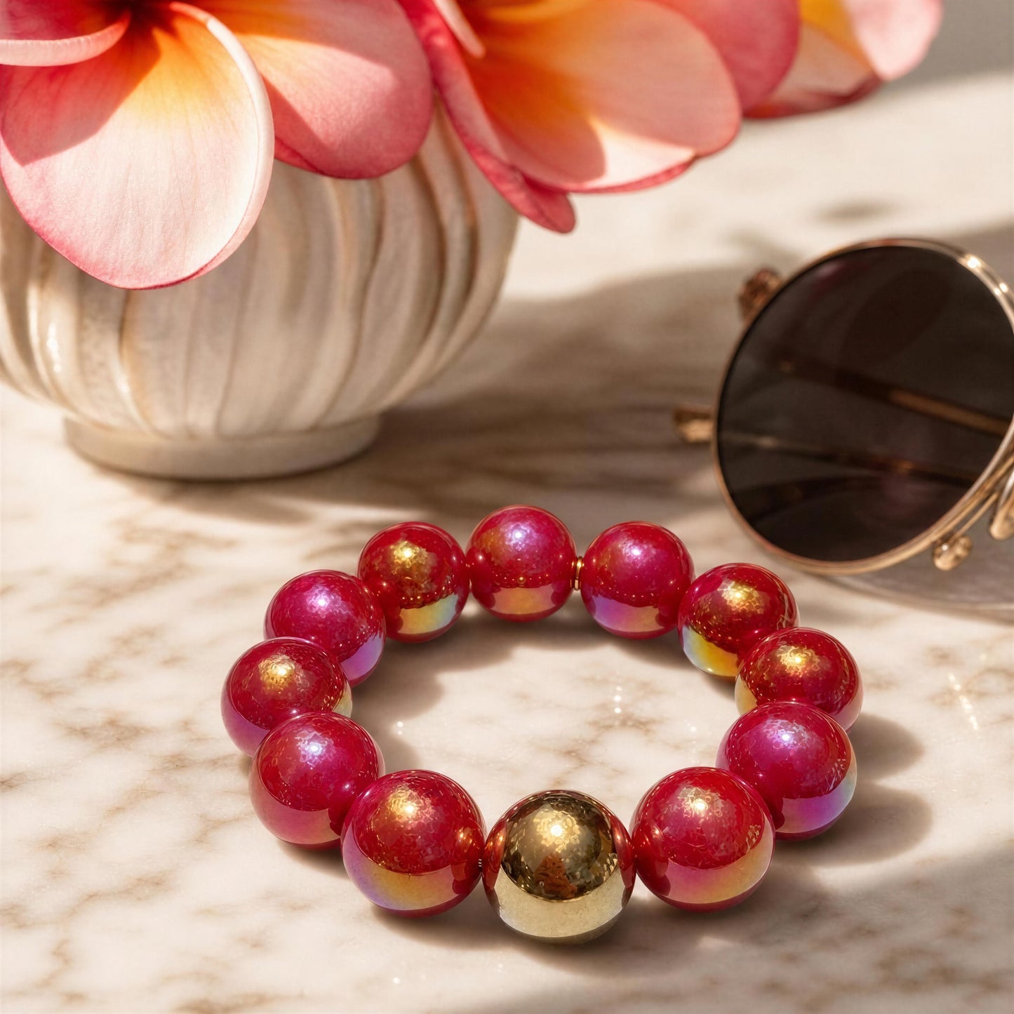 The Cherry Glow Bracelet with red and gold beads on a marble surface with flowers and sunglasses in the background