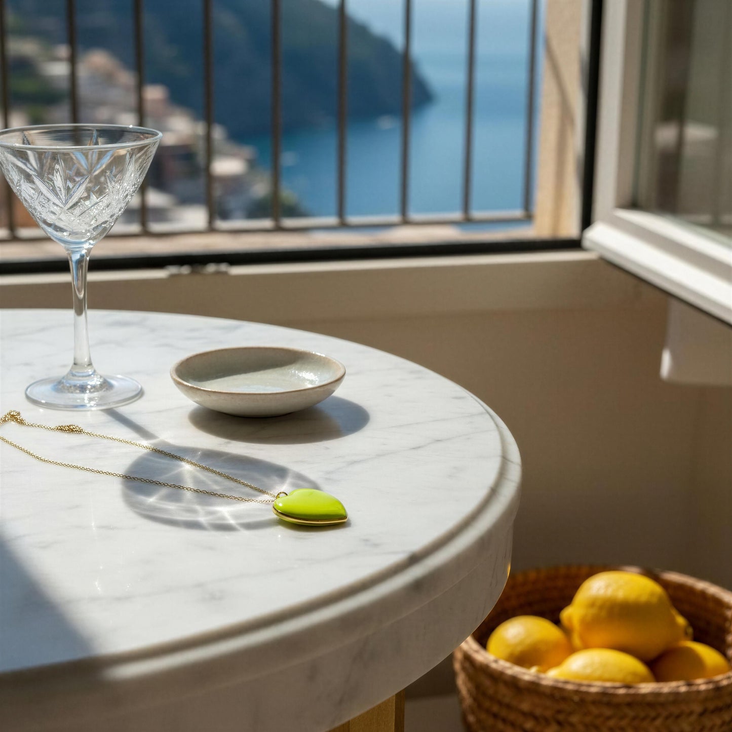 Round marble table with a glass, bowl, and basket of lemons in front of a window with a scenic view wearing the Limoncello Heart Necklace.