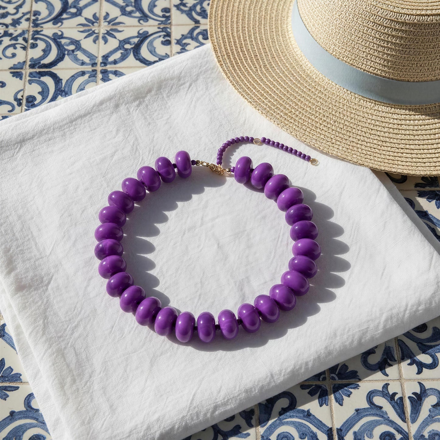 The Capri Lilla Choker arranged on a white fabric surface next to a straw hat on a blue-and-white patterned tile background.