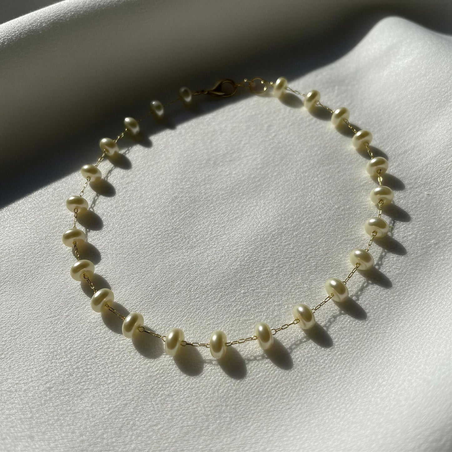 A delicate gold chain necklace adorned with small donut cream-colored freshwater pearls, displayed on a textured white fabric background under direct sunlight.