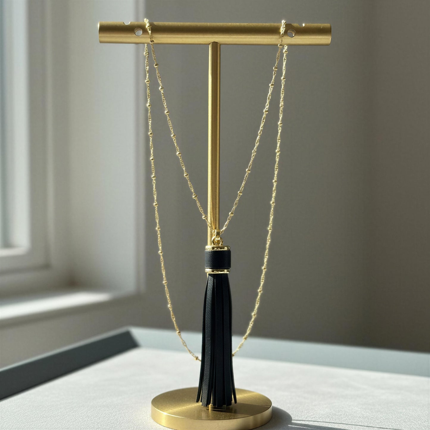 A gold T-stand jewelry display holder with a black tassel, holding the Frangia Ebano Necklace, set on a light gray surface with a soft-focus window background.