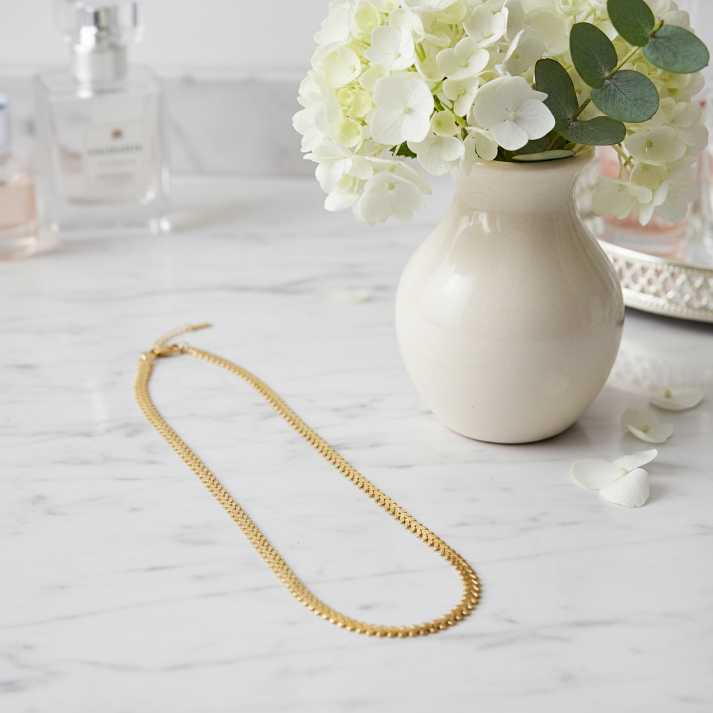 The Golden Vine Necklace on a marble surface with a vase of white flowers in the background