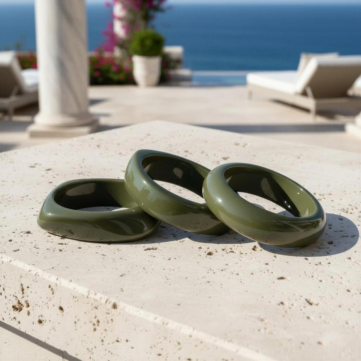 The Oliva Bangle Set from Chica Chica Jewelry . Three glossy olive green ceramic rings arranged on a light beige stone surface with an outdoor patio and ocean view in the background.