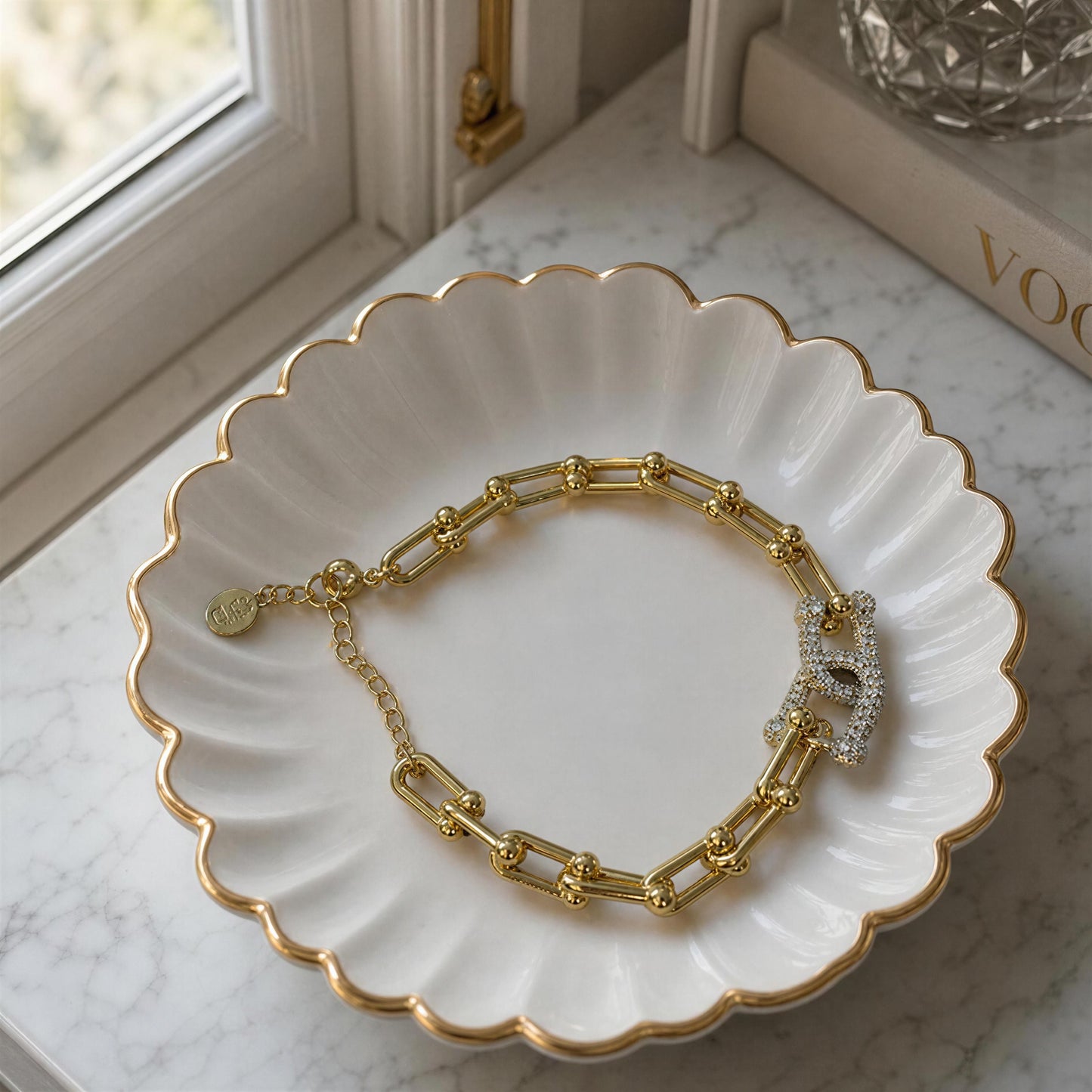 The Radiance Link Bracelet displayed on a white scalloped ceramic dish with gold trim on a marble surface near a window.