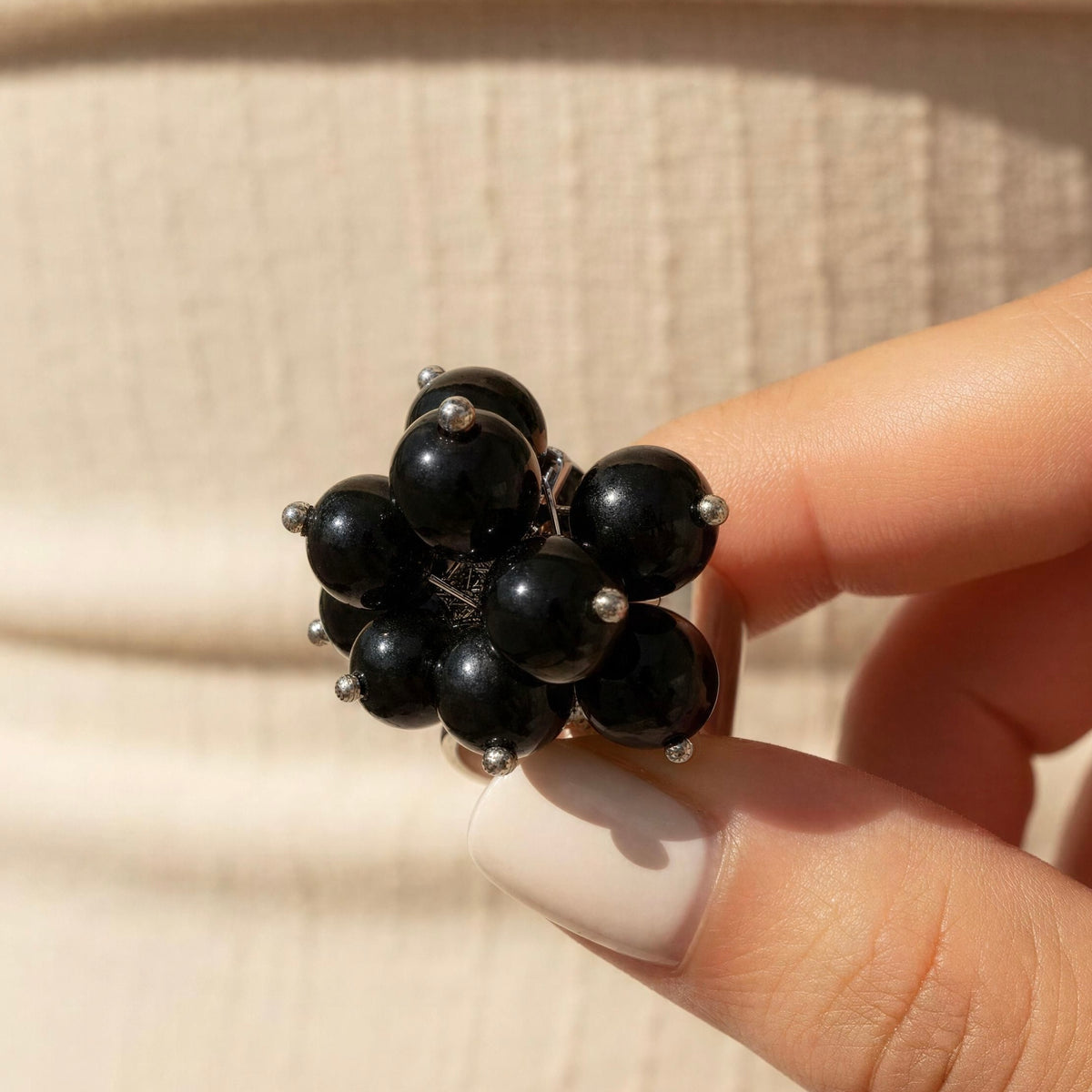 A close-up of a hand holding a cluster of glossy black spherical beads on a silver-toned ring, with a soft beige textured background.
