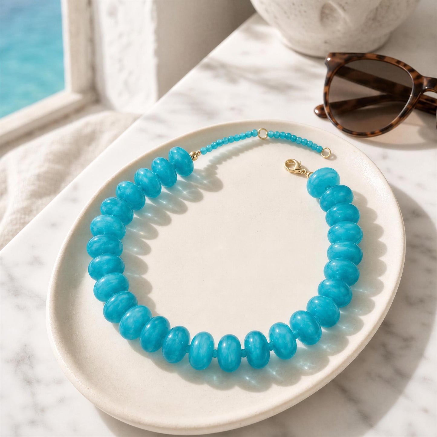 Turquoise beaded necklace on a white plate with sunglasses and a marble surface.