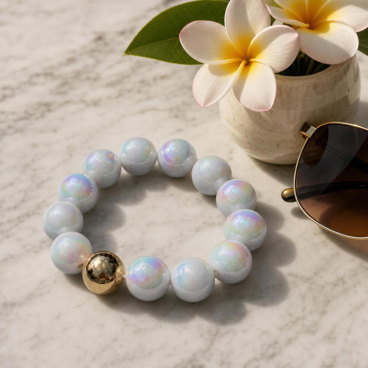 White acrylic beaded bracelet with a gold accent on a marble surface, next to a small potted plant and sunglasses.