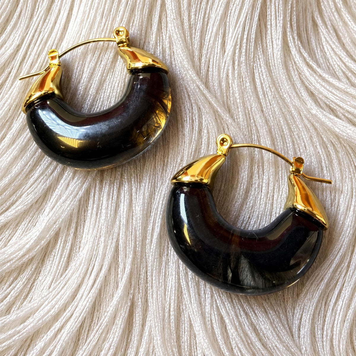 The Soraya Hoops Translucent Smoky Black Resin Earrings from Chica Chica Jewelry against white silk fringes fabric.