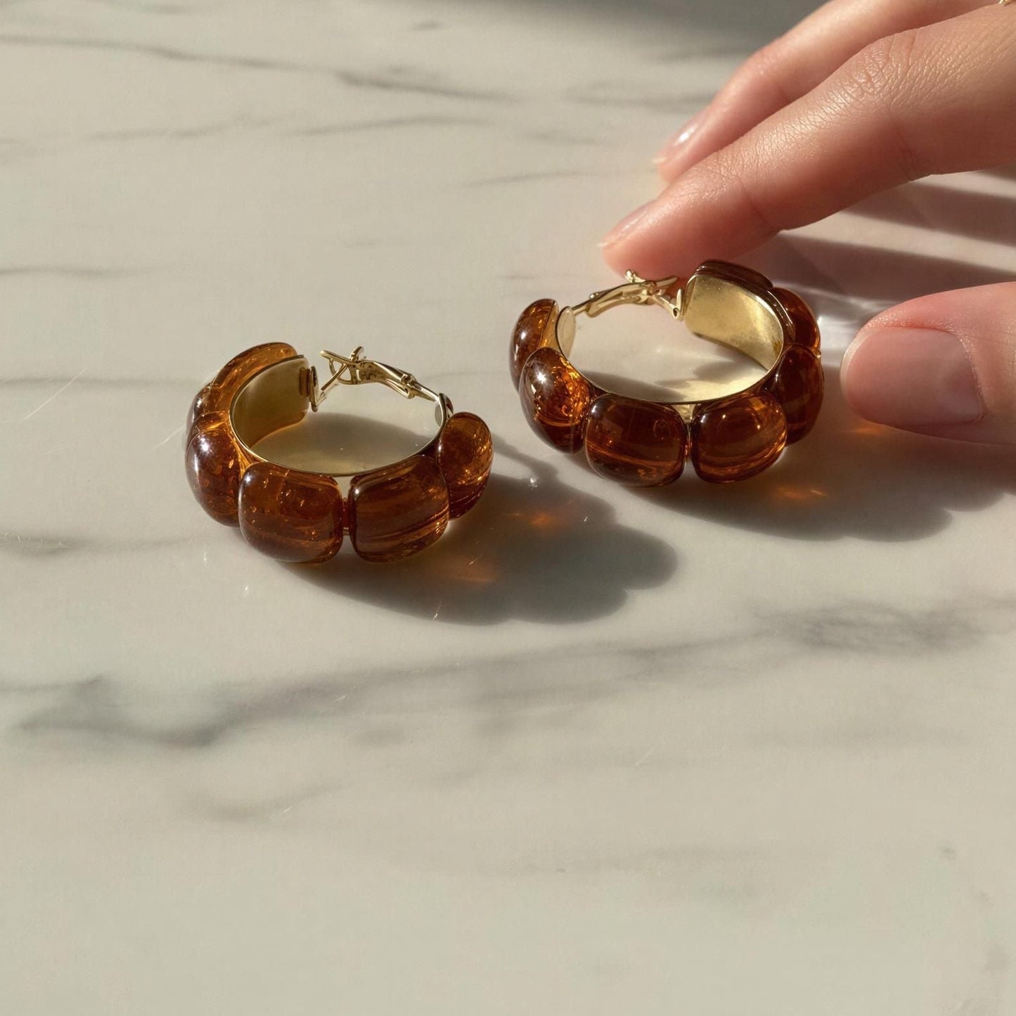 Hand holding a pair of amber-colored hoop earrings on a marble surface with a neutral background.