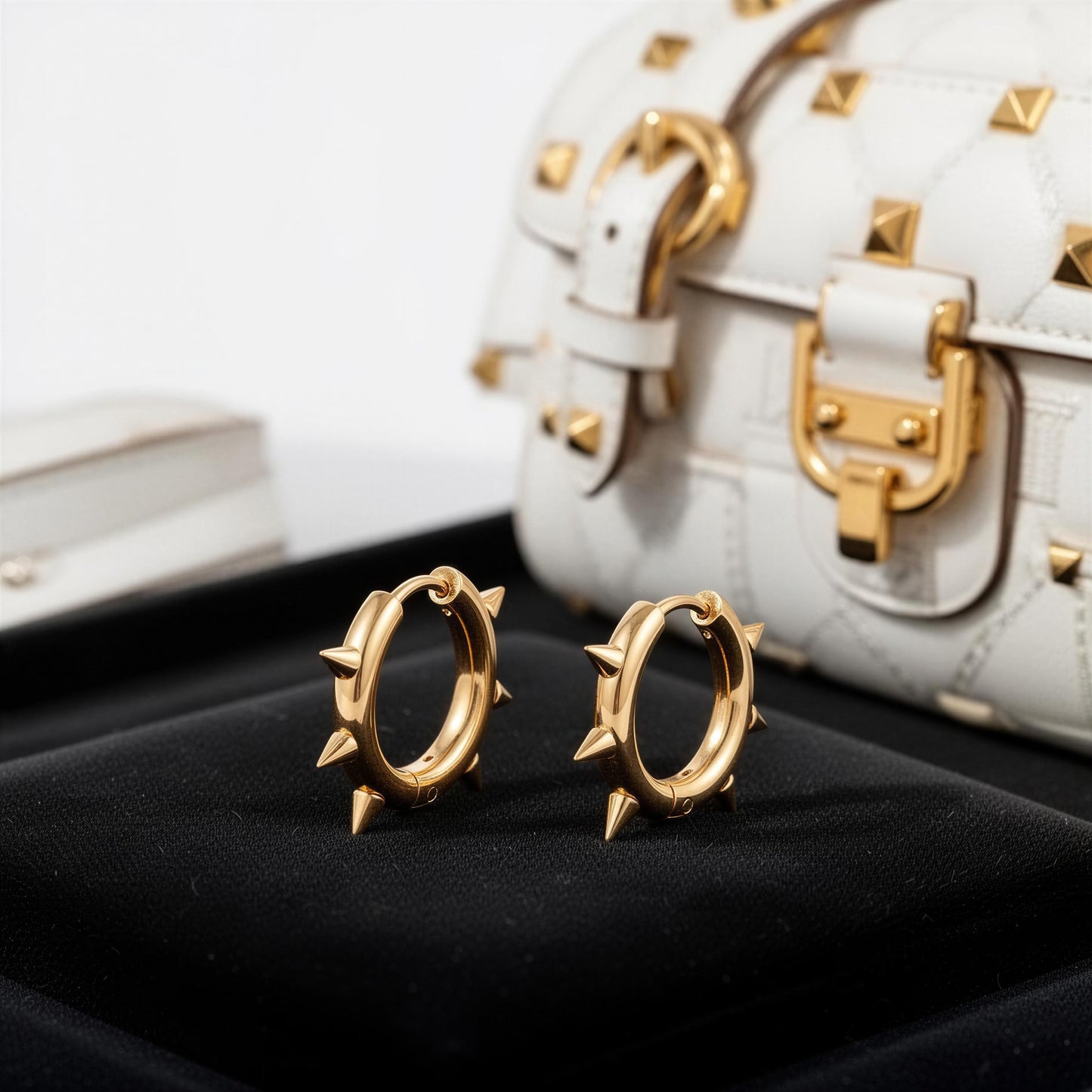 Gold hoop earrings with spike details on a black surface, with a white studded handbag in the background.