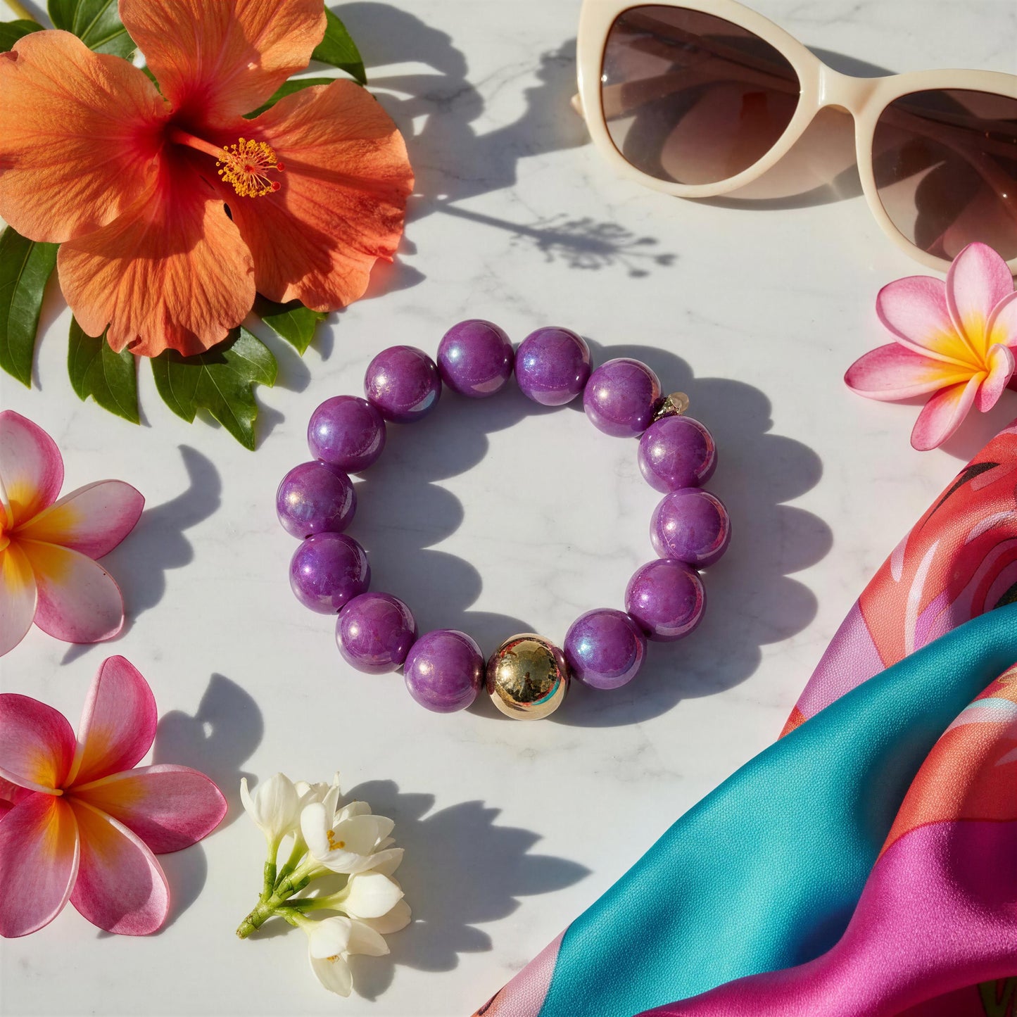 Purple beaded bracelet with a gold accent on a white surface surrounded by tropical flowers and sunglasses.