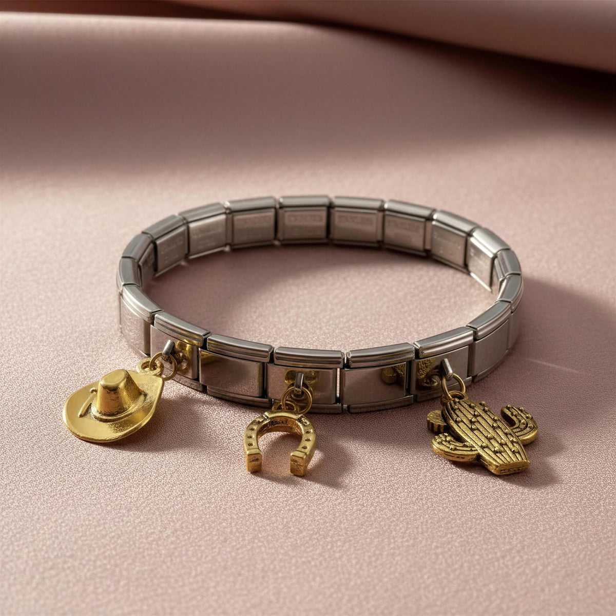 A silver-toned metal link bracelet with three dangling gold-tone charms, including a cowboy hat, a horseshoe, and a cactus, displayed on a soft pink fabric background.