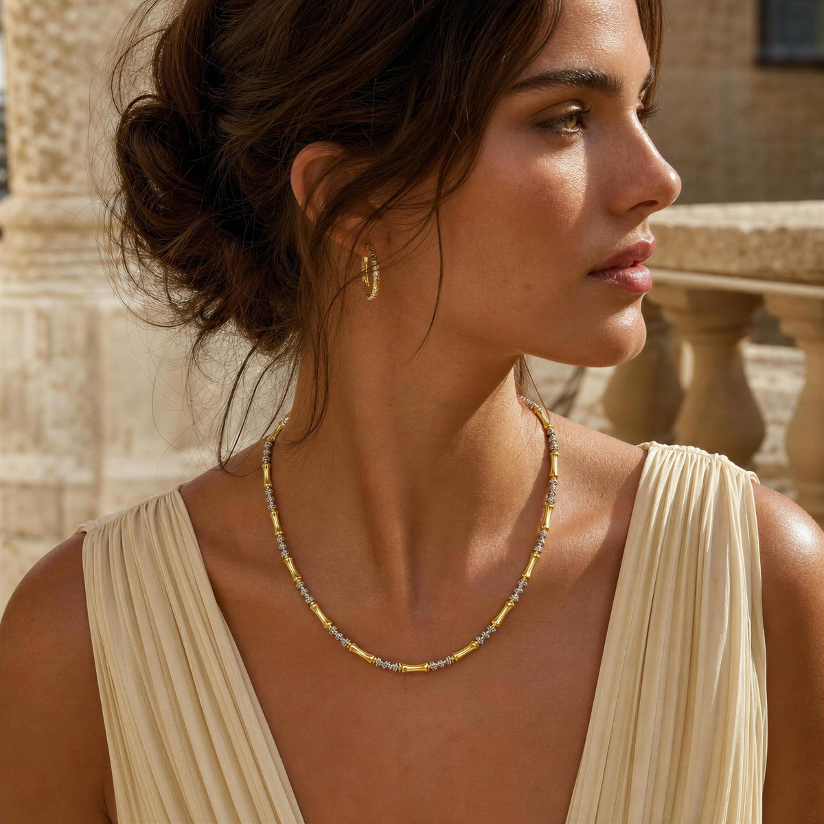 A woman in a cream pleated dress wearing the Malta Necklace from Chica Chica Jewelry, a delicate necklace, photographed outdoors against a balustrade.