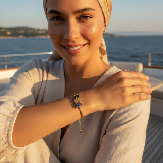 A woman wearing the Aegea Bracelet from Chica Chica Jewelry , a gold chain bracelet with, blue, champagne color and green crystal-embellished charm, posing on a boat deck with the sea in the background.