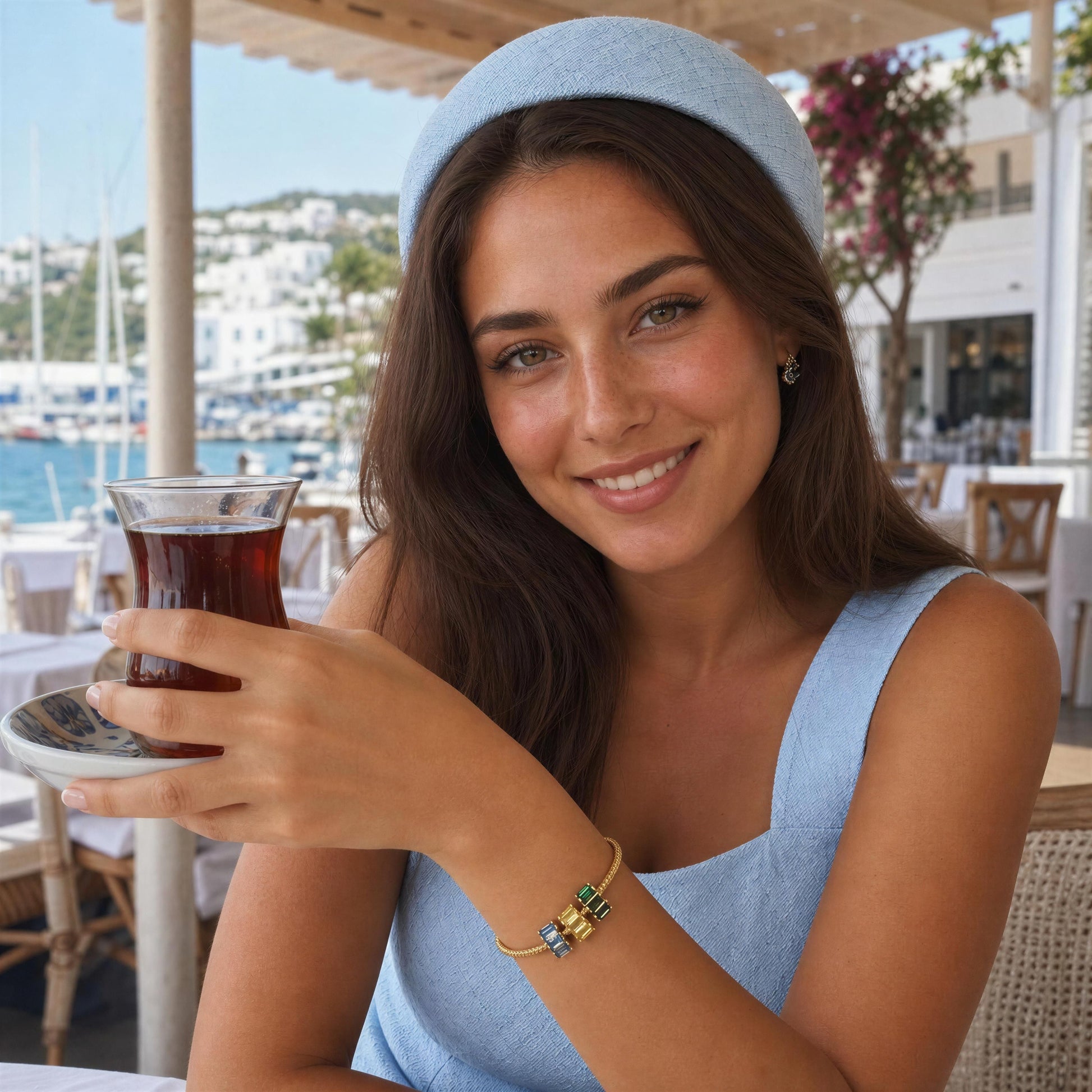 A woman wearing a light blue headband and matching top holds a glass of tea at an outdoor cafe, wearing a gold charms bracelet on her wrist.