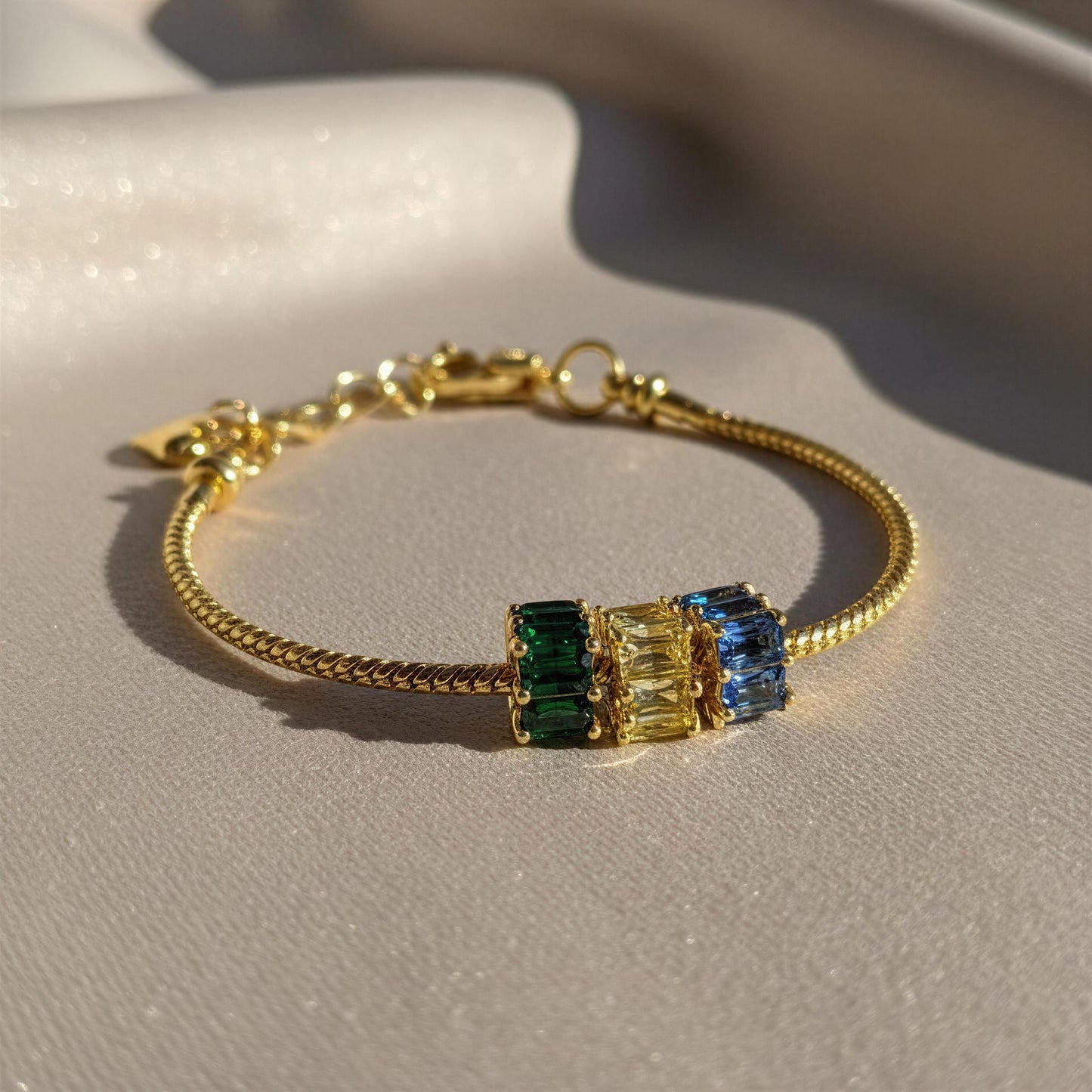 A gold-plated snake chain bracelet with three rectangular bezel-set crystal charms in green, yellow, and blue, displayed on a textured light beige surface.
