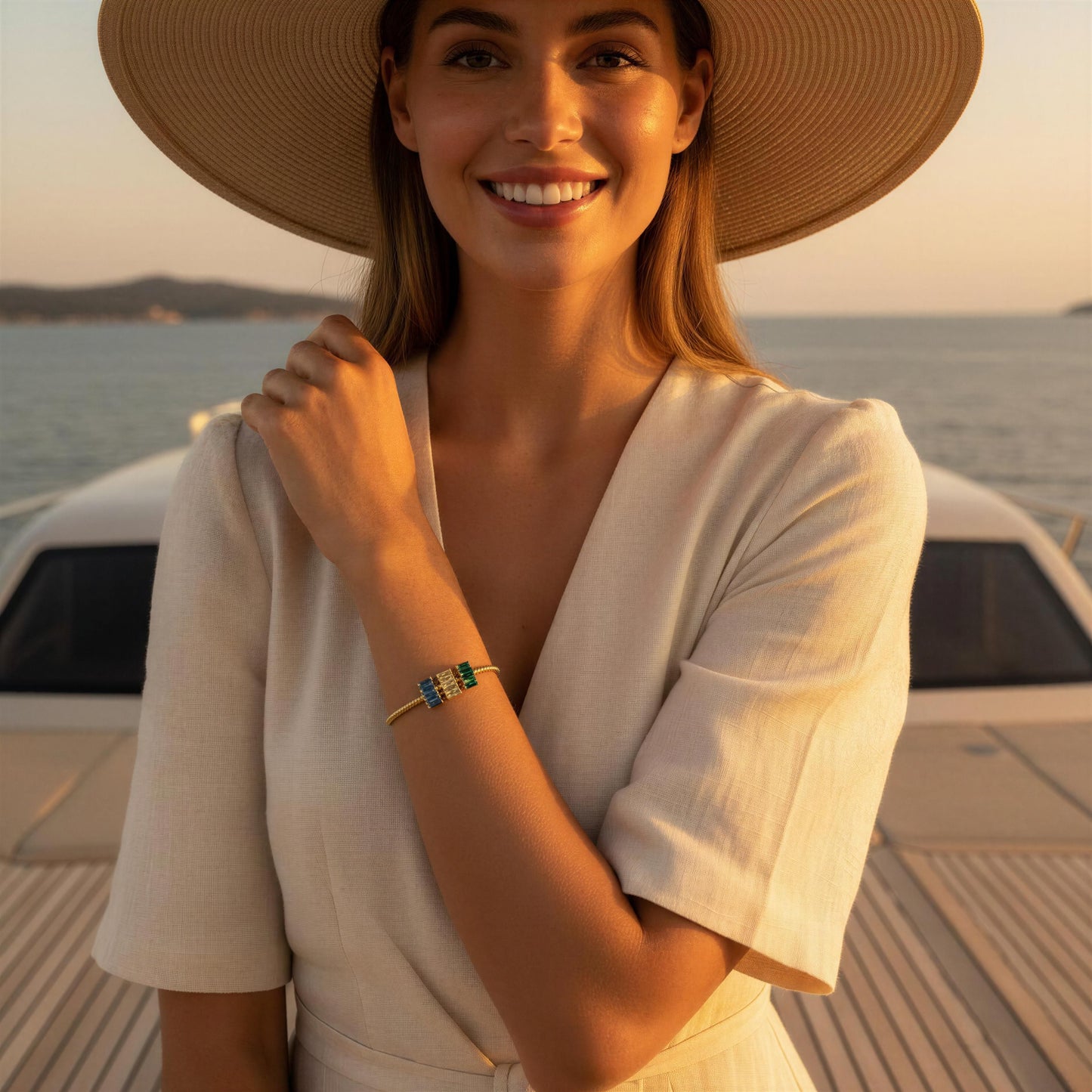 A woman wearing a wide-brimmed straw hat and a cream-colored short-sleeve top stands on a boat deck at sunset, wearing a delicate gold bracelet with green and blue stones.