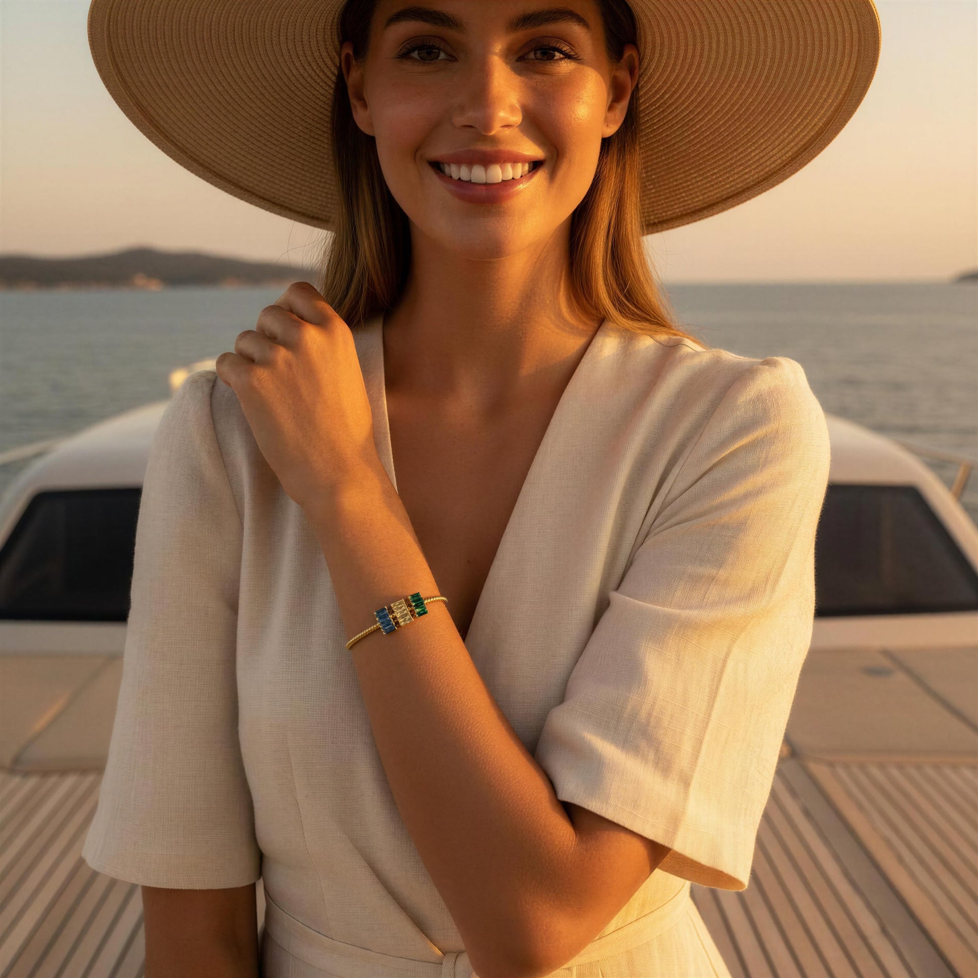A woman wearing a wide-brimmed straw hat and a cream-colored short-sleeve top stands on a boat deck at sunset, wearing a delicate gold bracelet with green and blue stones.