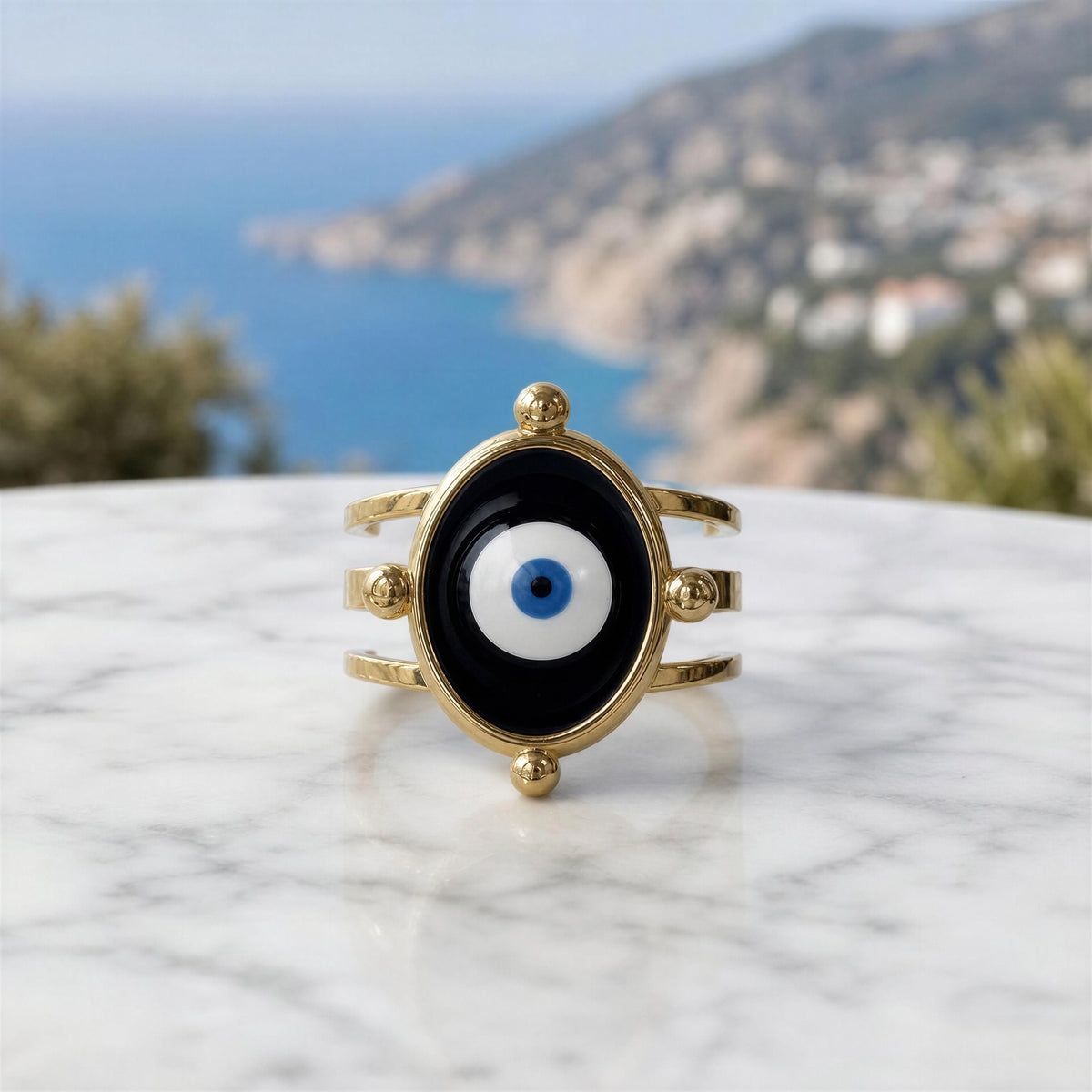 A gold-tone evil eye ring with a black oval face and blue-and-white center, displayed on a white marble surface with a blurred coastal landscape in the background.
