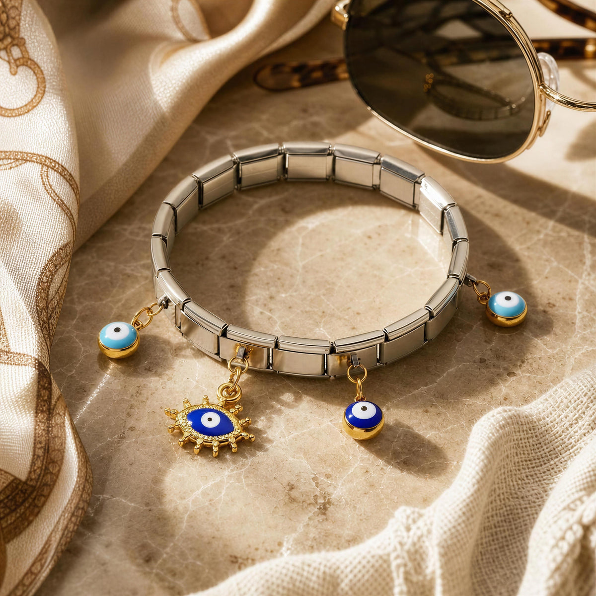 A silver-toned segmented bracelet with three evil eye charms, styled on a beige marble surface with draped cream fabric and gold-framed sunglasses in the background.