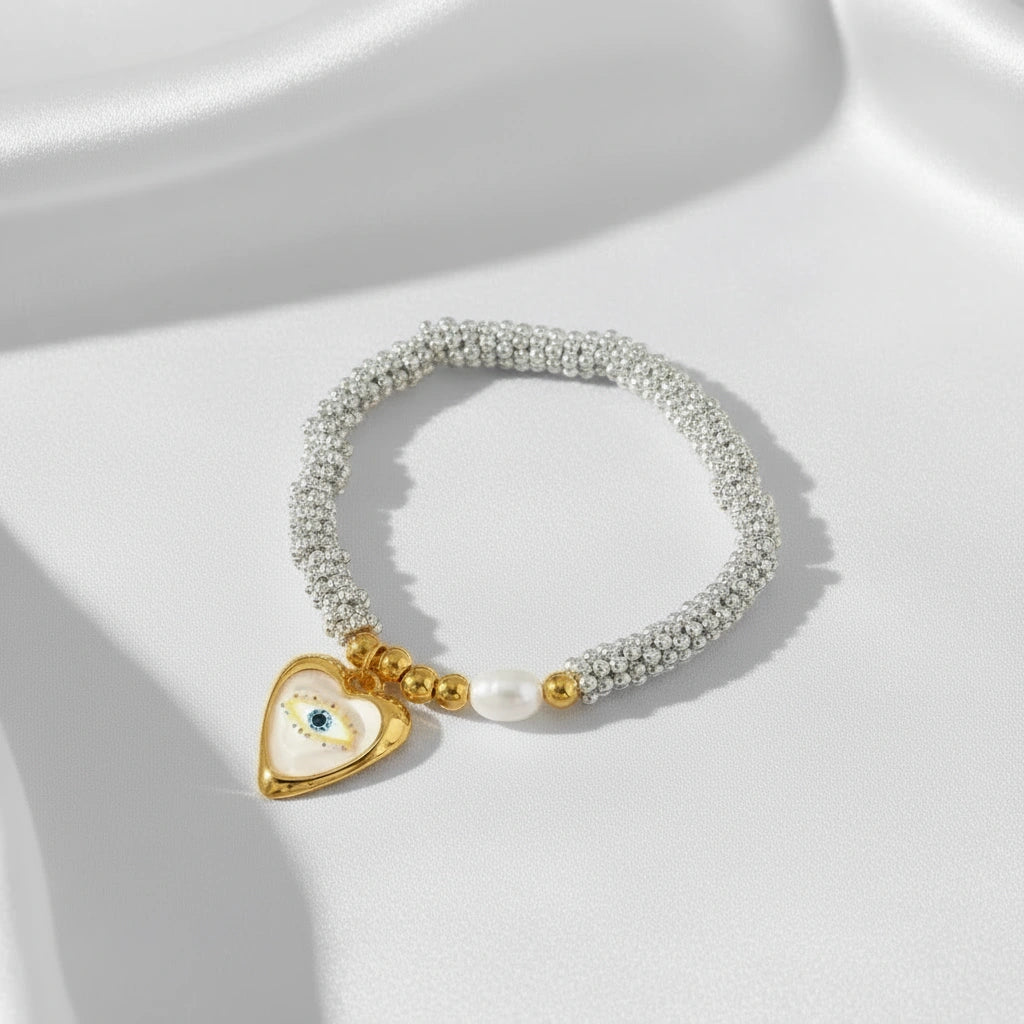 Aegis Aura silver beaded stretch bracelet with gold accents, freshwater pearl and heart-shaped evil eye charm on a clean white background.