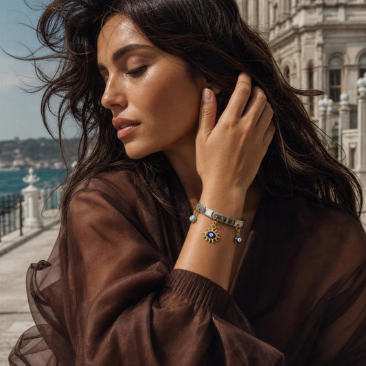A woman wearing the Nazar Bracelet from  Chica Chica Jewelry  a silver-toned bracelet with blue and gold details, photographed outdoors near a waterfront and historic building.