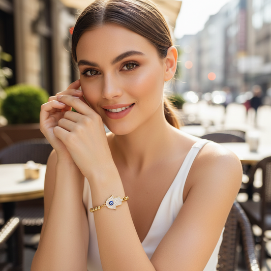 Model wearing the Aegis Guardian Hand Bracelet with pavé hamsa charm and blue evil eye, styled at an outdoor café.
