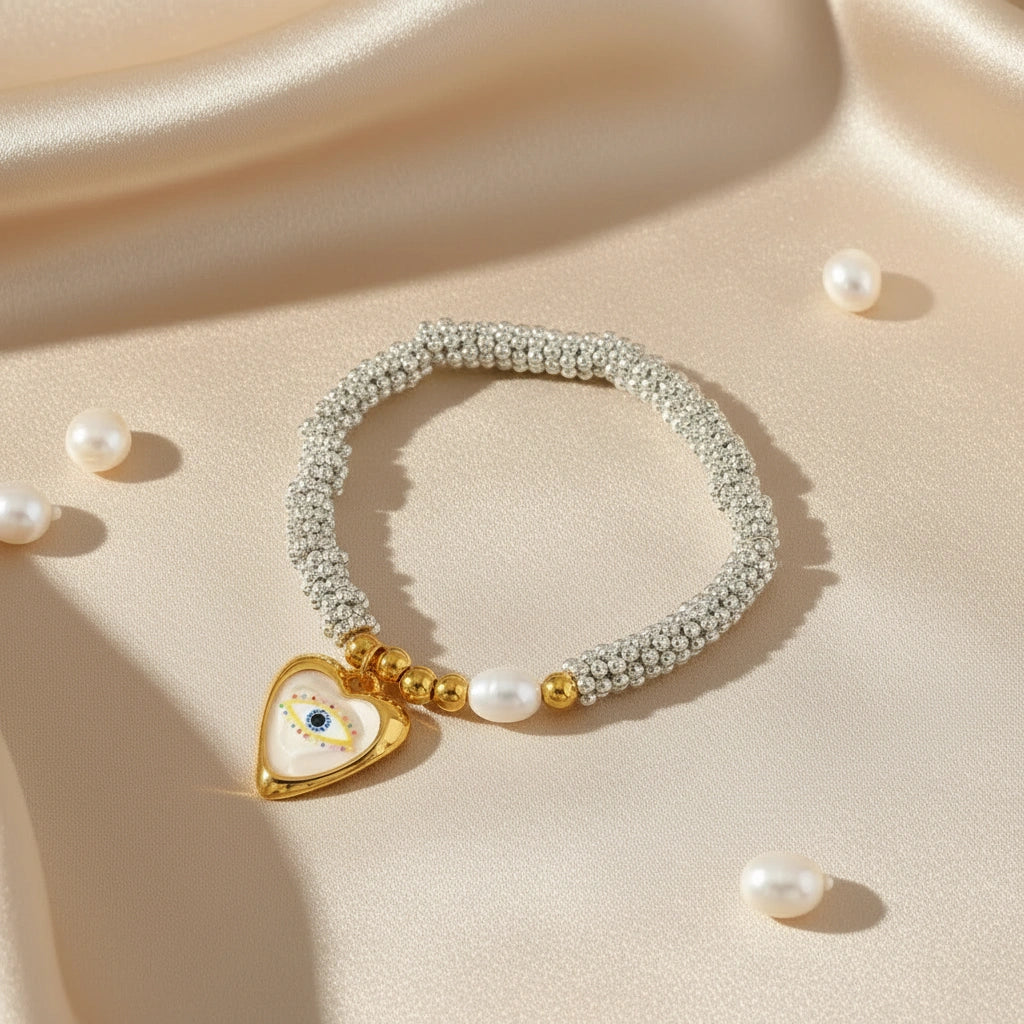 The Luma bracelet flatlay on soft satin fabric with pearls, showcasing silver beaded stretch bracelet, gold details and heart evil eye charm.