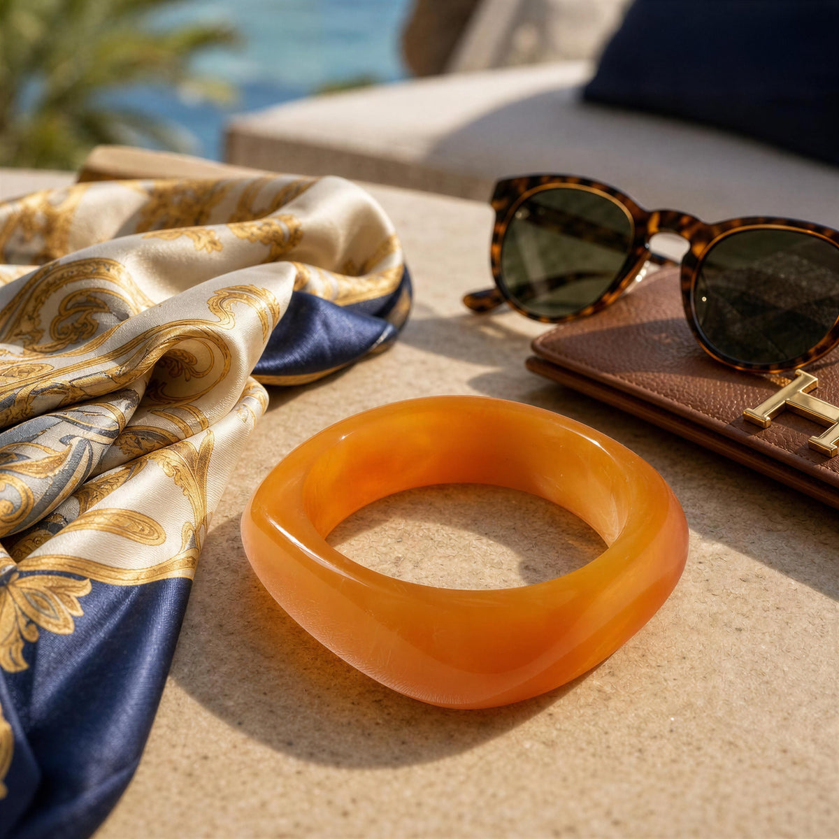 The Alev Bangle from Chica Chica Jewelry; a translucent orange bangle bracelet displayed on a beige stone surface next to a gold and navy silk scarf, tortoiseshell sunglasses, and a brown leather wallet.