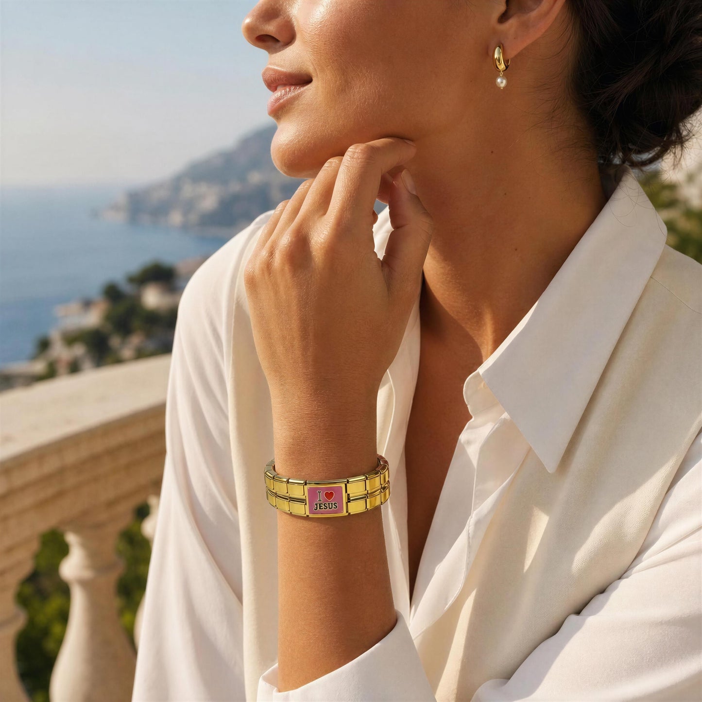 A woman in a white collared shirt wearing a gold-plated bracelet with pink enamel and a central heart-shaped logo, photographed outdoors with a coastal background.
