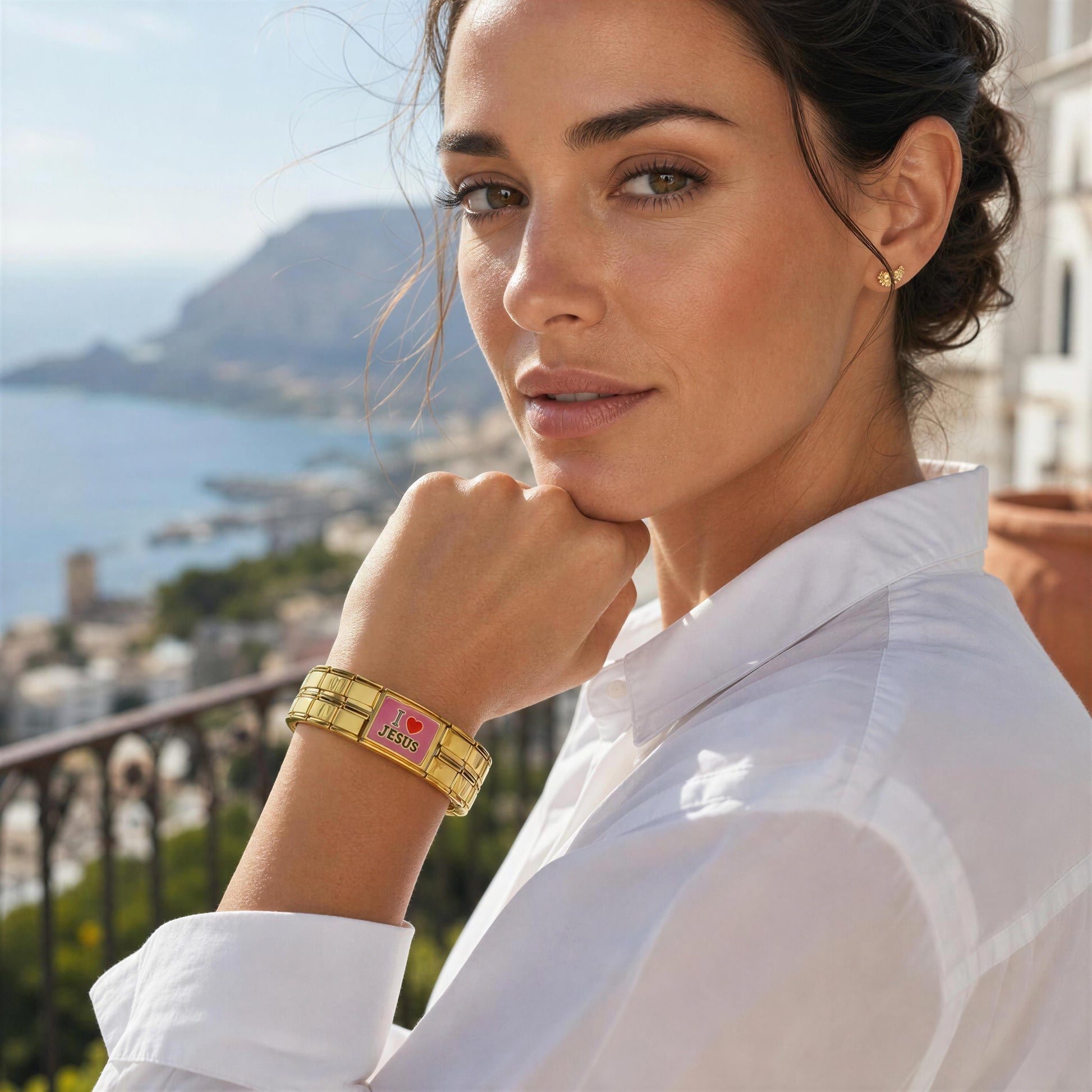 A woman in a white shirt wearing a gold-tone bangle bracelet with the Amada Bracelet from Chica Chica Jewelry, posed outdoors with a coastal city and mountain background.