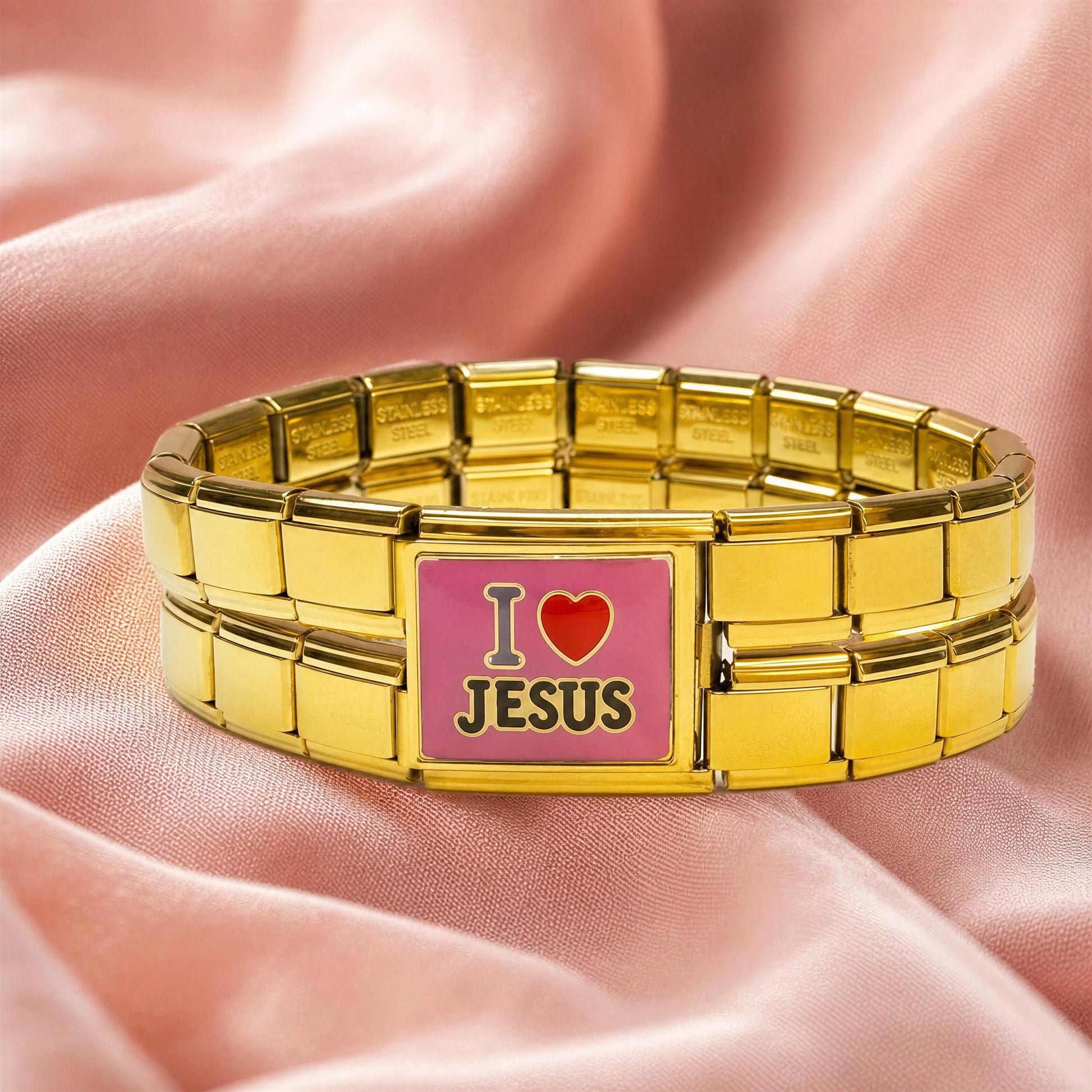 A gold-tone Italian link bracelet with a rectangular center plate that reads 'I love JESUS' in black text on a pink background, displayed on draped pink satin fabric.