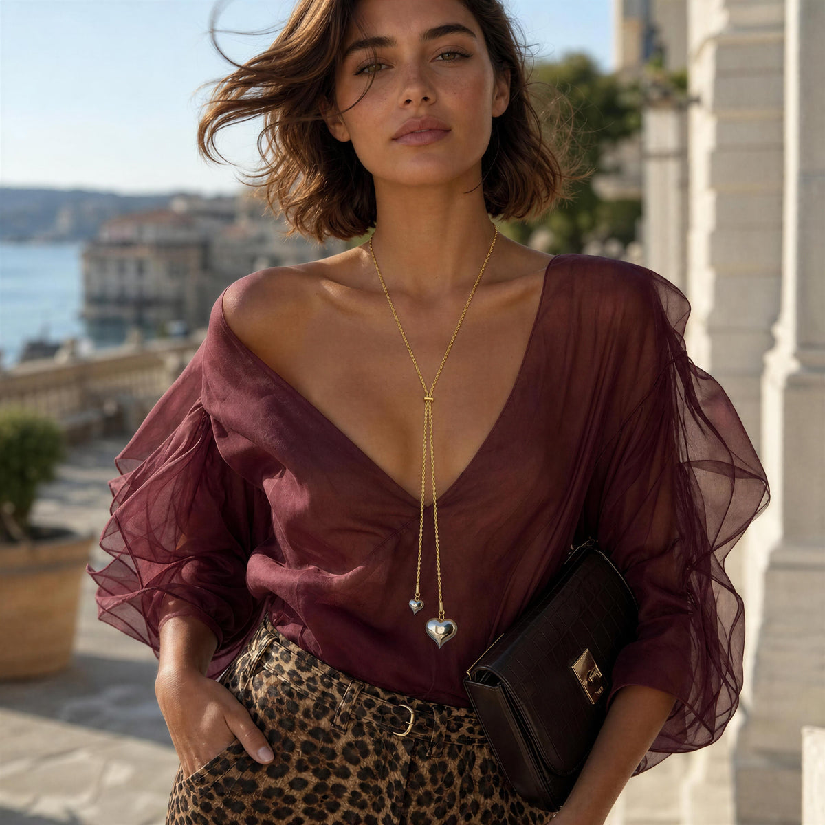 A woman wearing a sheer burgundy off-the-shoulder blouse with dramatic ruffled sleeves, paired with leopard-print pants and a long gold necklace with a heart pendant, holding a dark brown clutch bag in an outdoor coastal setting.
