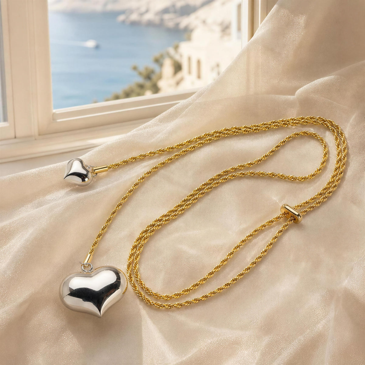 A gold and silver heart pendant necklace draped on cream-colored satin fabric, with a window showing a scenic view in the background.
