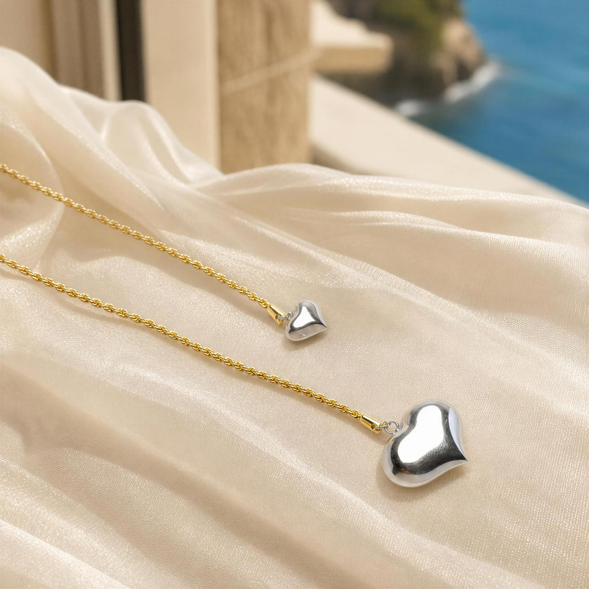 Two heart-shaped pendant necklaces, one gold and one silver, draped on cream-colored satin fabric with a blurred coastal background.