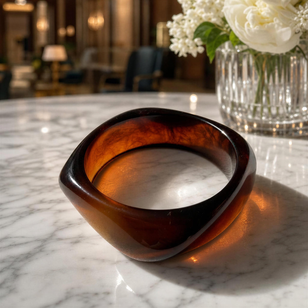 The Ambra Bangle
from Chica Chica Jewelry; a thick, chunky translucent amber-brown glass bowl with a rounded, organic shape, resting on a white marble tabletop with a blurred background of a modern interior and white flowers in a vase.