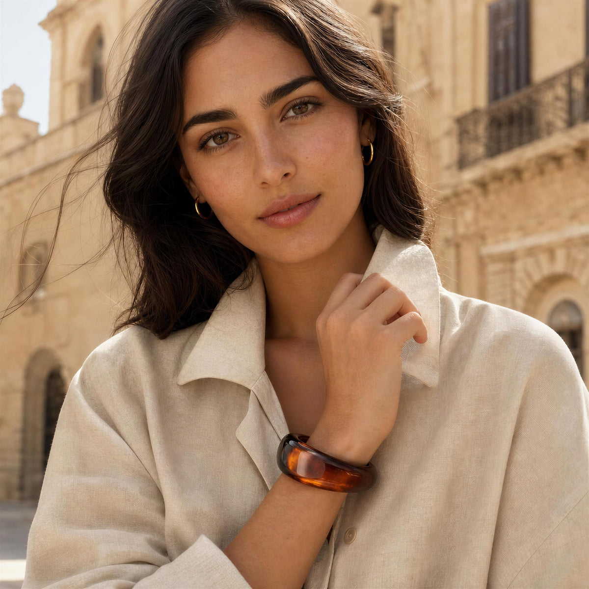 A woman wearing the Ambra Bangle
From Chica Chica Jewelry, a tortoiseshell bangle watch and small gold hoop earrings, posing outdoors in front of a warm beige stone building.