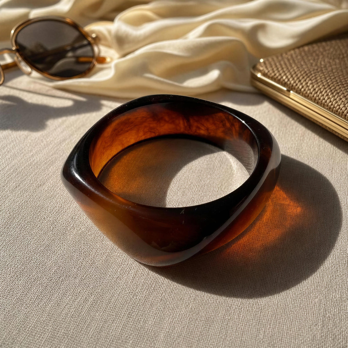 The Ambra Bangle from Chica Chica Jewelry. A thick, chunky resin bangle with a glossy dark amber-brown tortoiseshell pattern, styled on a beige linen fabric with draped cream silk, gold-framed sunglasses, and a woven clutch.