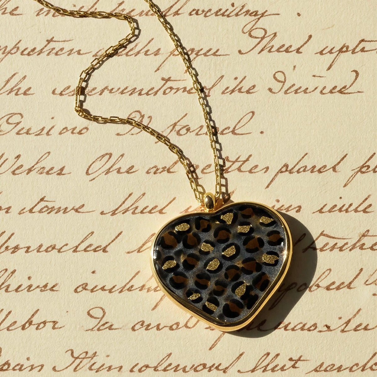A heart-shaped pendant necklace with a leopard print pattern, featuring black and brown spots with gold glitter accents, set in a gold-tone frame on a gold chain. The pendant is displayed on a vintage-style beige paper background with cursive writing.