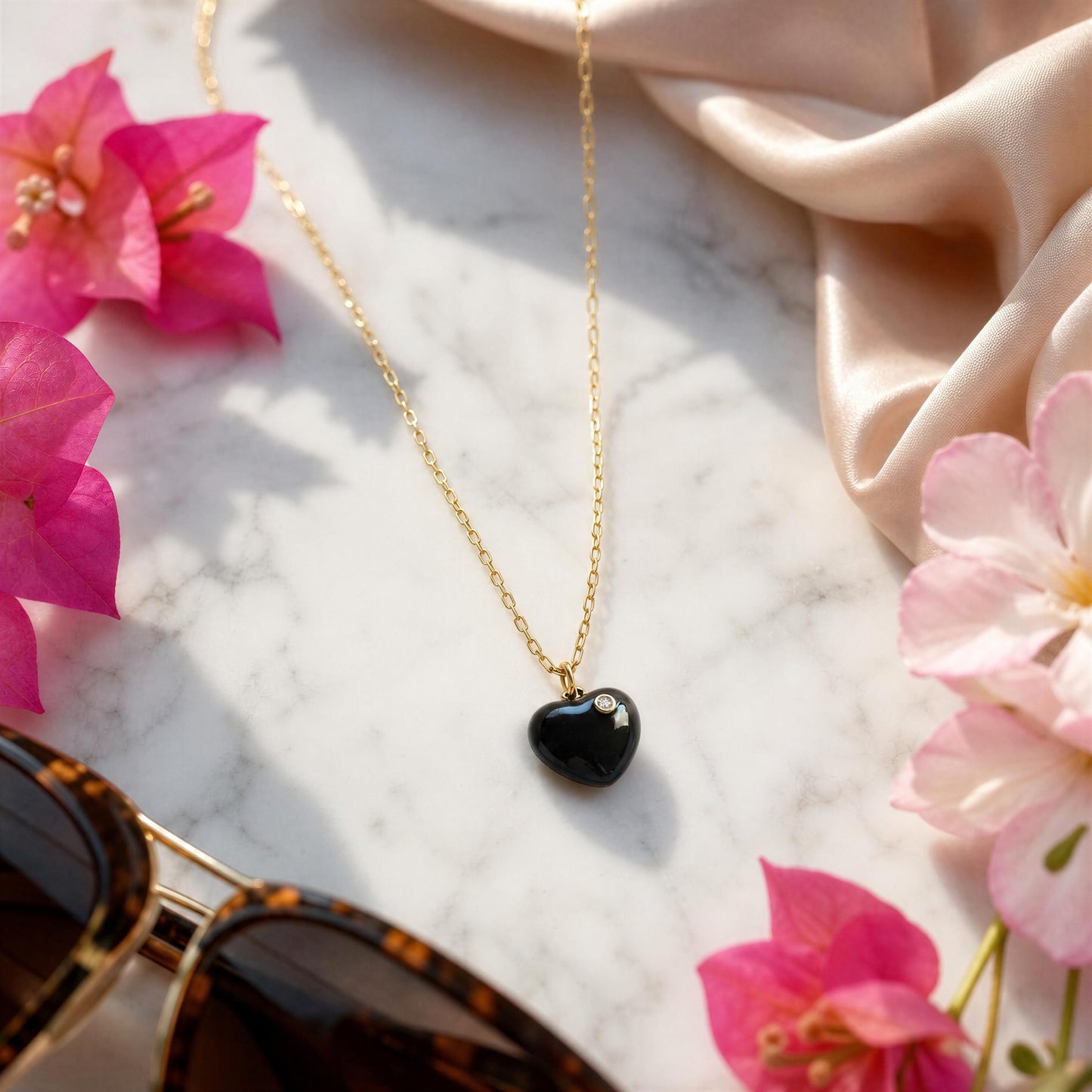 Amore Noir Heart Necklace on a marble surface with pink flowers and sunglasses.