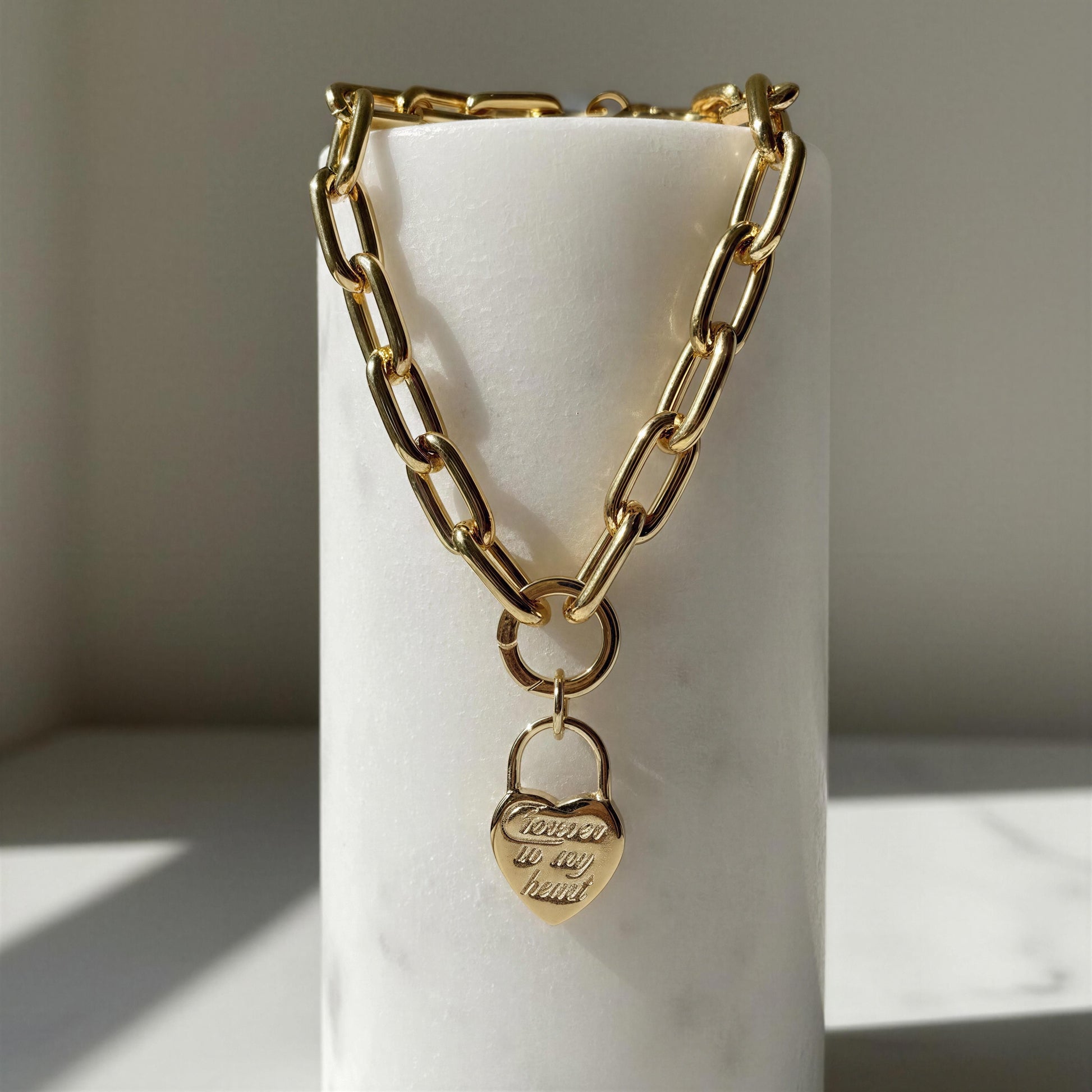 Gold chain necklace with the Amore Lock Necklace - a heart-shaped pendant on a white cylindrical stand.