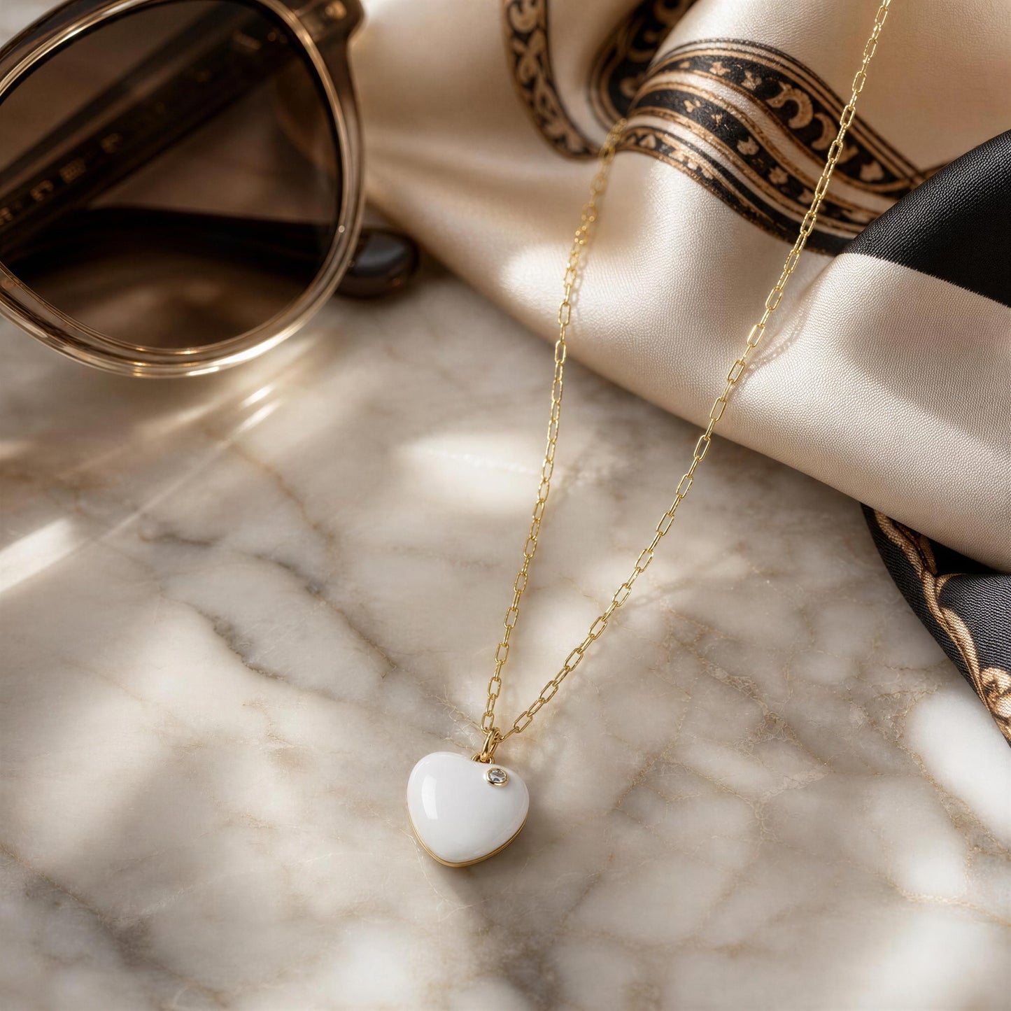 The Amore Moonlight Heart Necklace on a marble table next to sunglasses and headscarf.