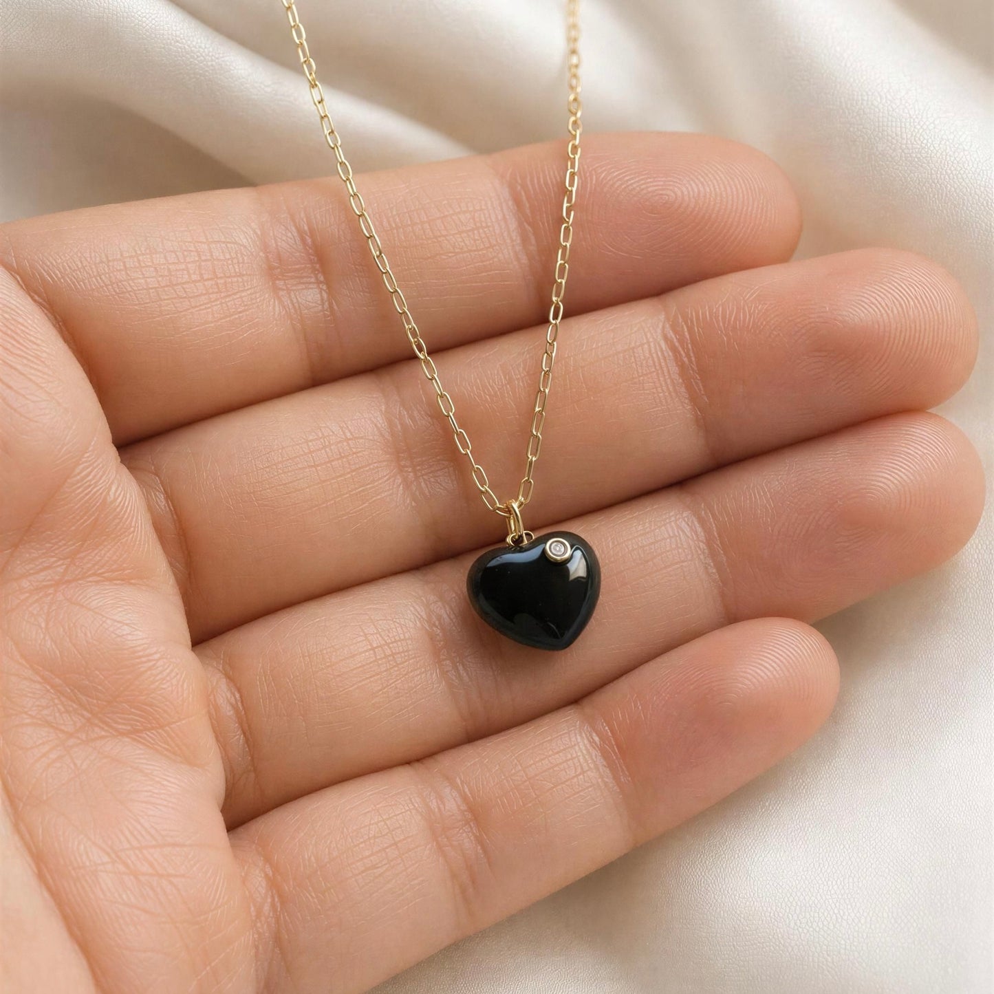 Black heart-shaped pendant on a gold chain held in a hand against a white background