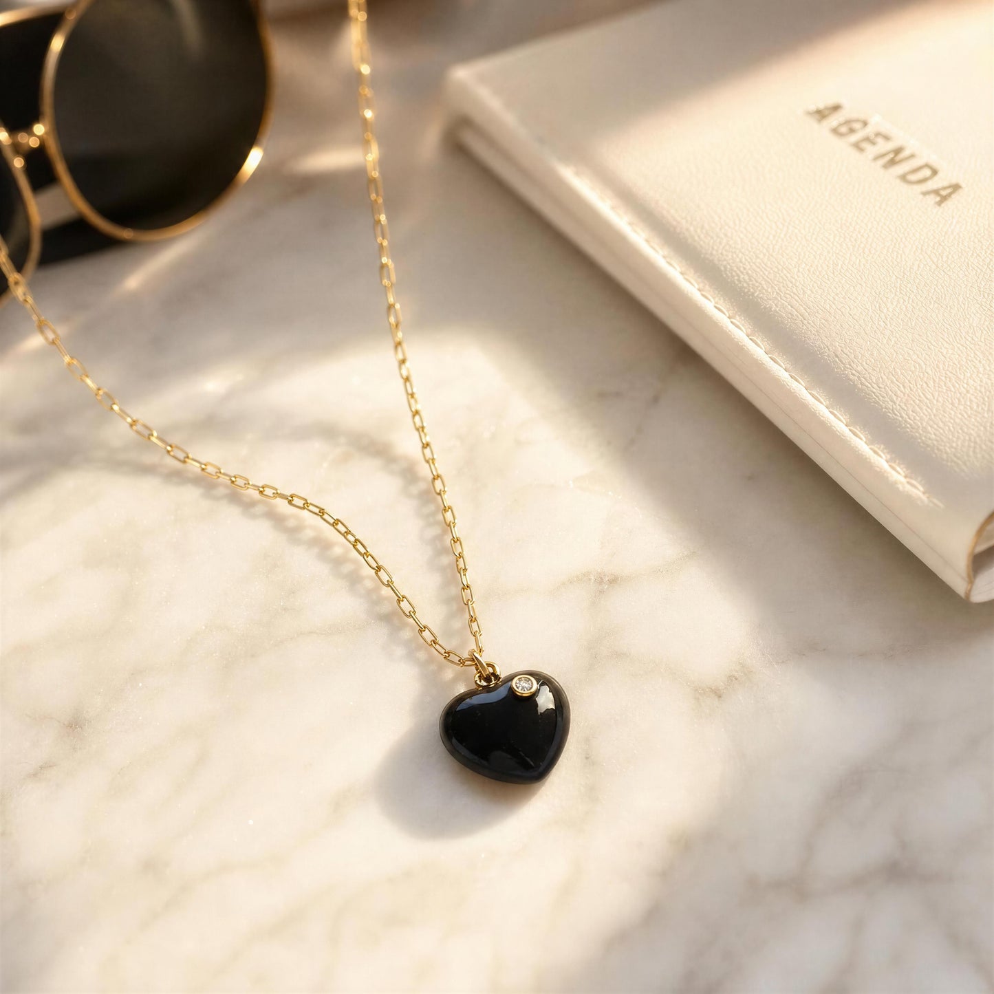 The Amore Noir Heart Necklace on a marble surface next to sunglasses and an agenda.