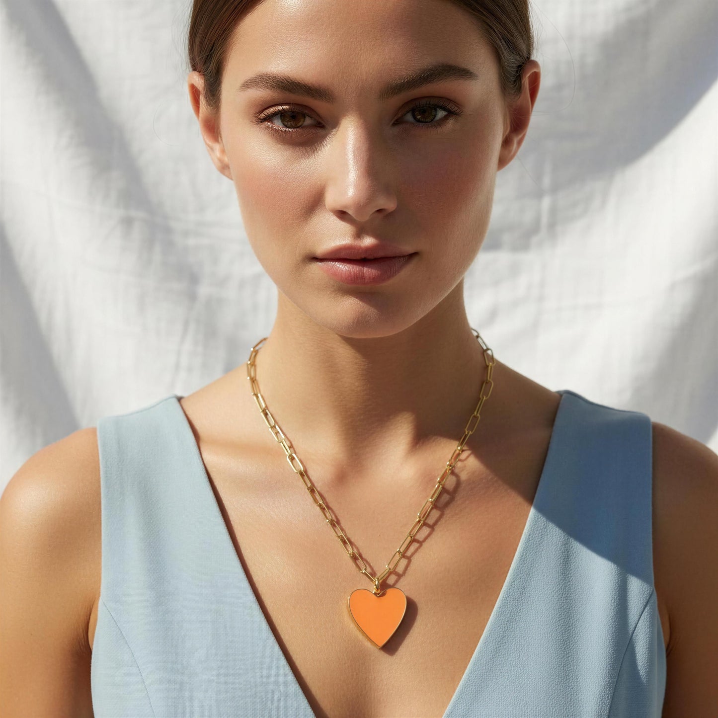 Woman wearing a light blue sleeveless top and a gold chain necklace with an orange heart pendant against a white background.