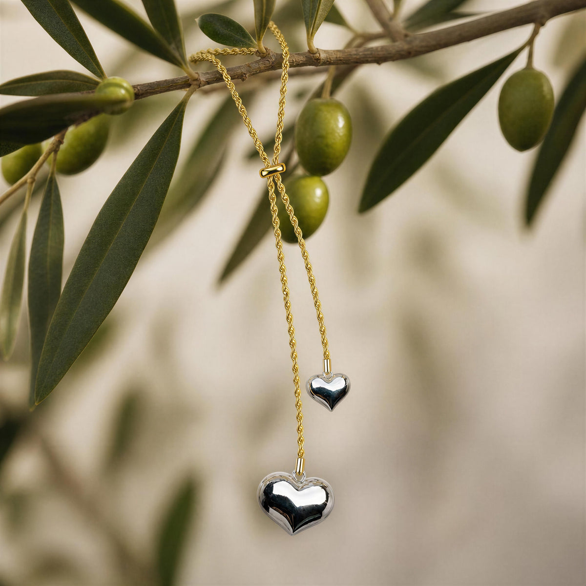 A gold-tone lariat-style necklace with two dangling heart charms, one matte gold and one polished silver, hanging from an olive branch with green olives against a soft beige background.